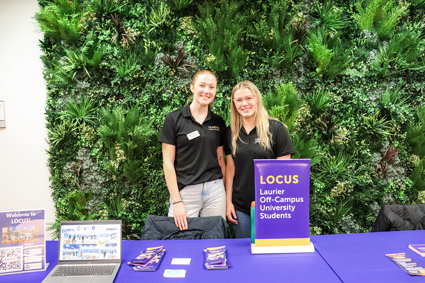 Two volunteers standing behind a table in with a "Laurier Off-Campus University Students" sign, brochures and a laptop