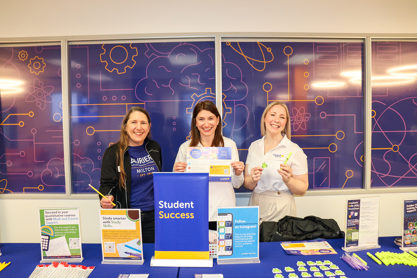 Three volunteers standing behind a table with signage about Student Success services, stickers and highlighters