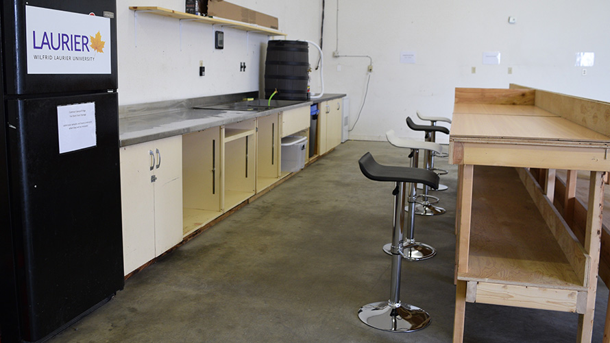 A lab with four stools facing a wooden bench, opposite a counter with cupboard space next to a fridge.