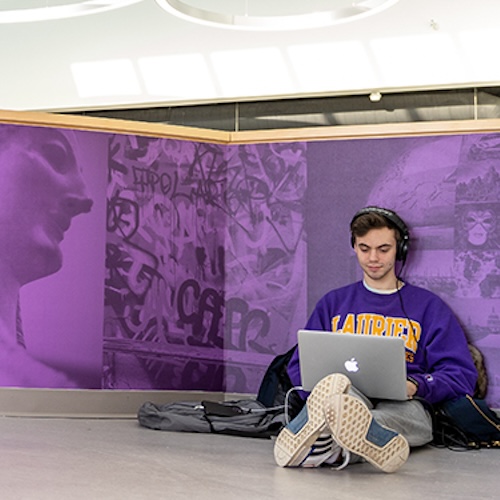 Student sitting on the floor working on a laptop with headphones on, leaning against the wall of a balcony with Laurier branding