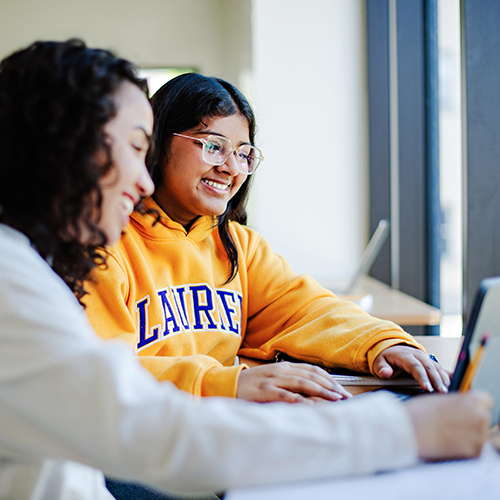 Two students sitting at a desk looking at tablet screen and taking notes