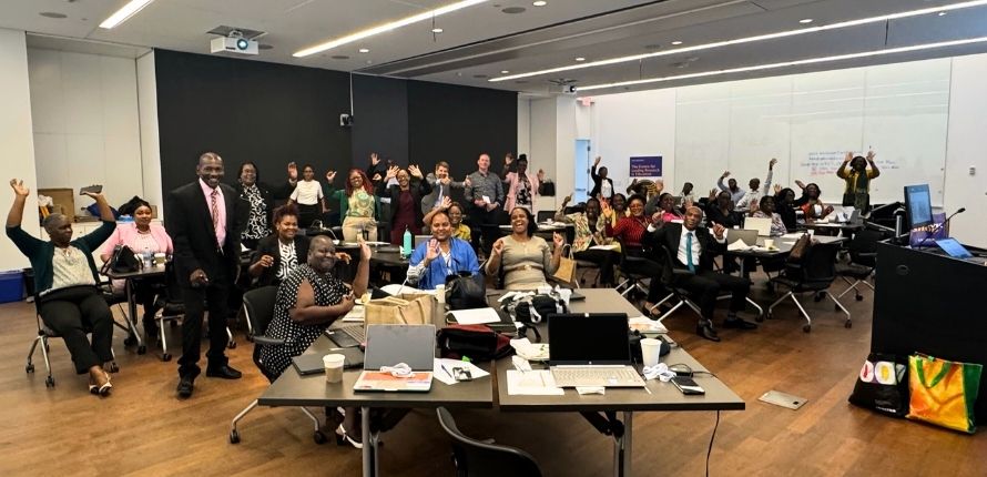A group of people in a conference room wave at the camera.