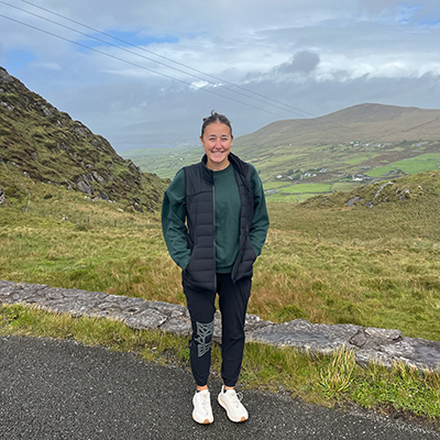 A young woman stands in front of a rugged Irish landscape